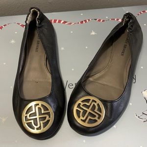 Audrey Brooke black flats with gold emblem.
